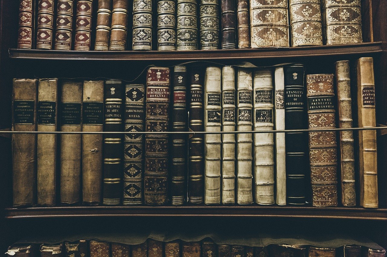A close up of a bookshelf with antique and leather bound books on it, mostly in creams, browns, and blacks. The titles are illegible.